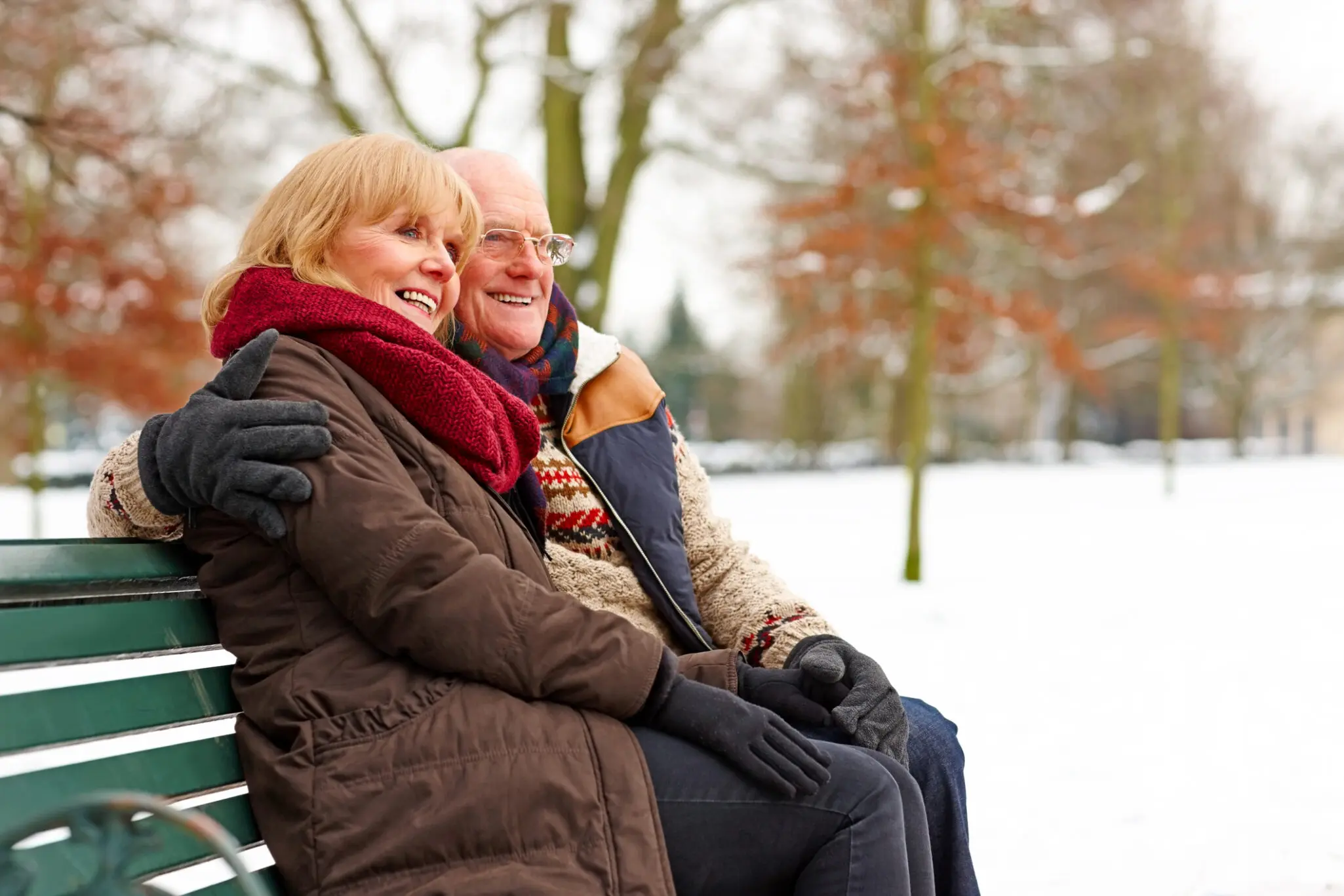 Elderly couple sitting on a bench at snow park senior companionship: older couple wearing heavy jackets and scarves snuggles on bench in the snow